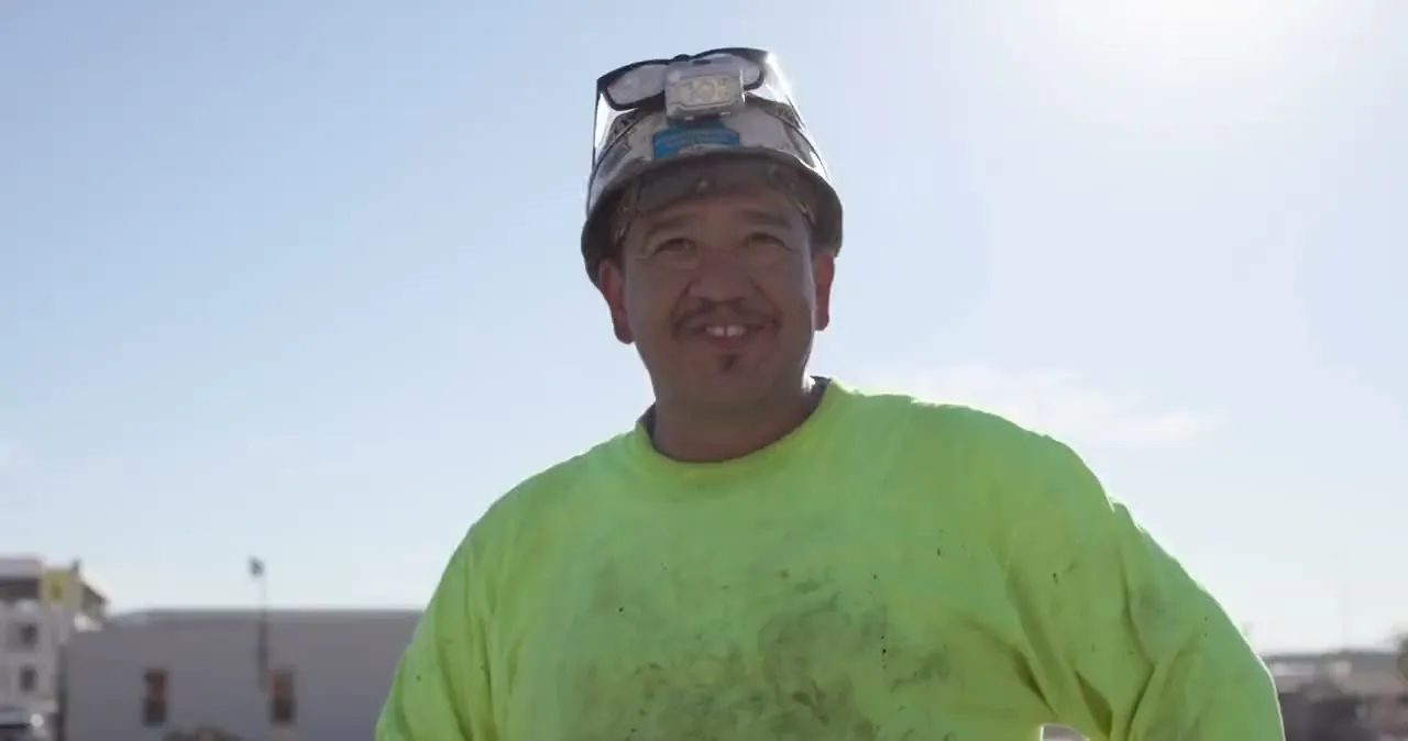 An ironworker in a hard hat and a hi-vis shirt, smiling, against a clear sky