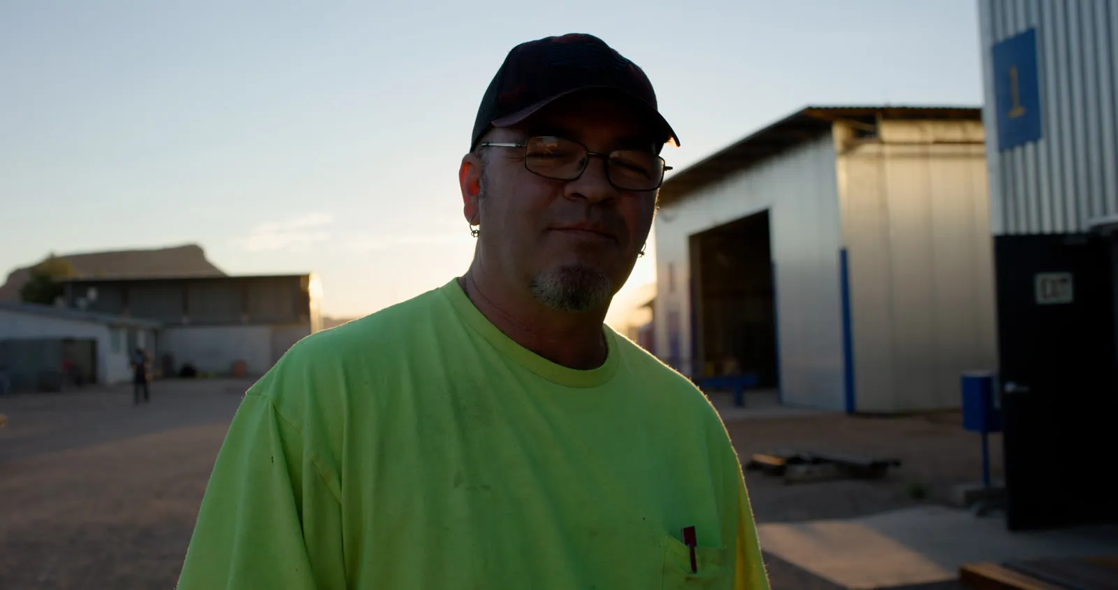 Fabricator in a neon-yellow shirt, cap and glasses standing in a steel shop yard at sunset
