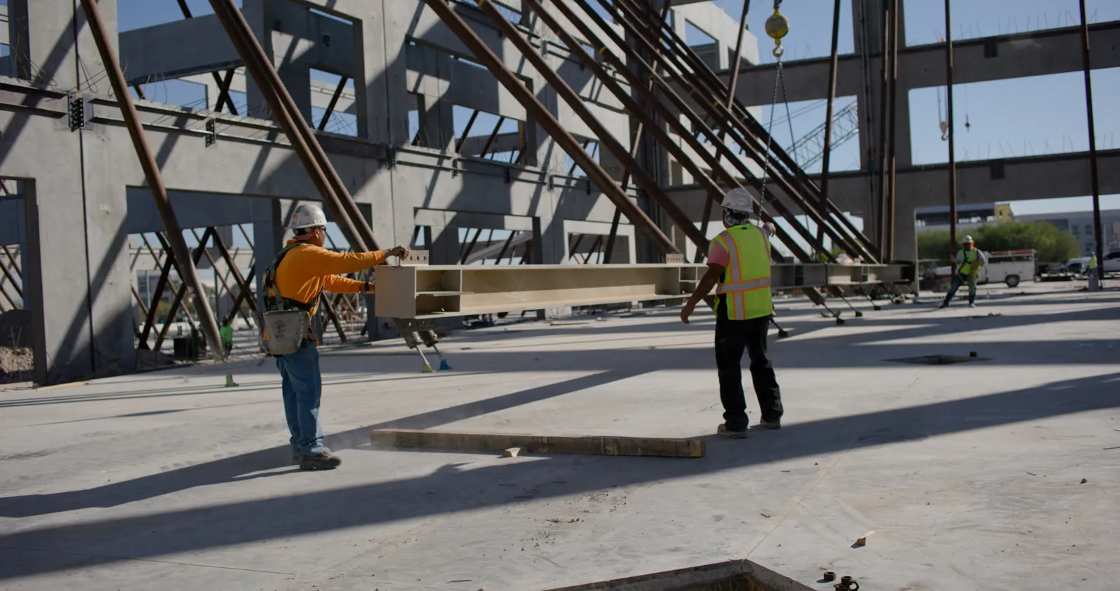 Two ironworkers in hard hats and hi-vis guiding a steel beam on a crane hook across the slab, between braced precast walls