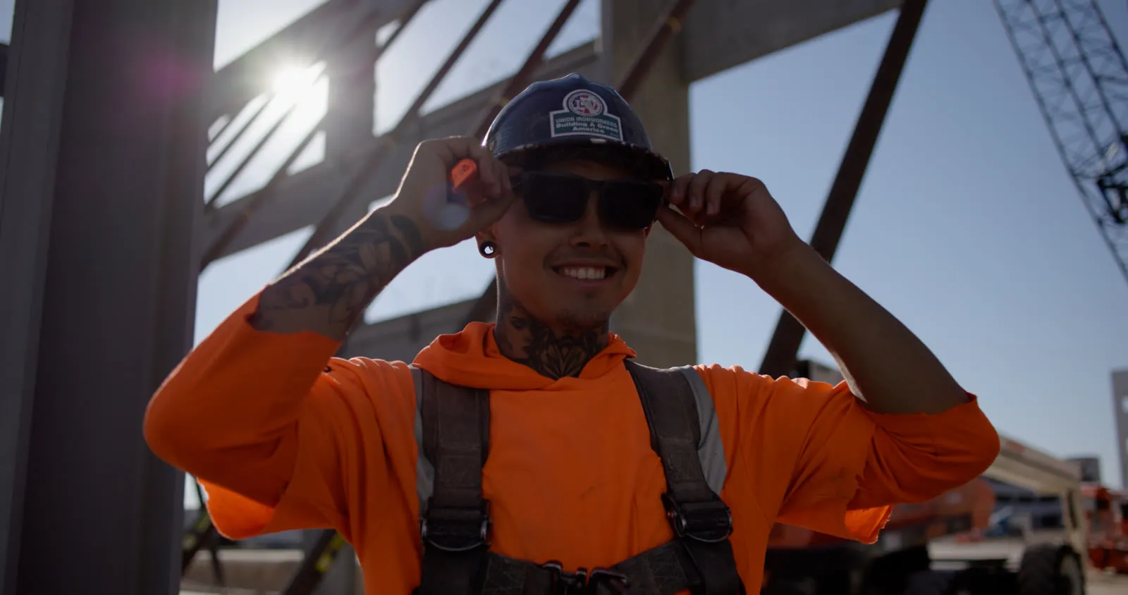 Ironworker in an orange hoodie, harness and hard hat smiling as he adjusts his sunglasses on a job site, a precast and steel frame and a crane behind him