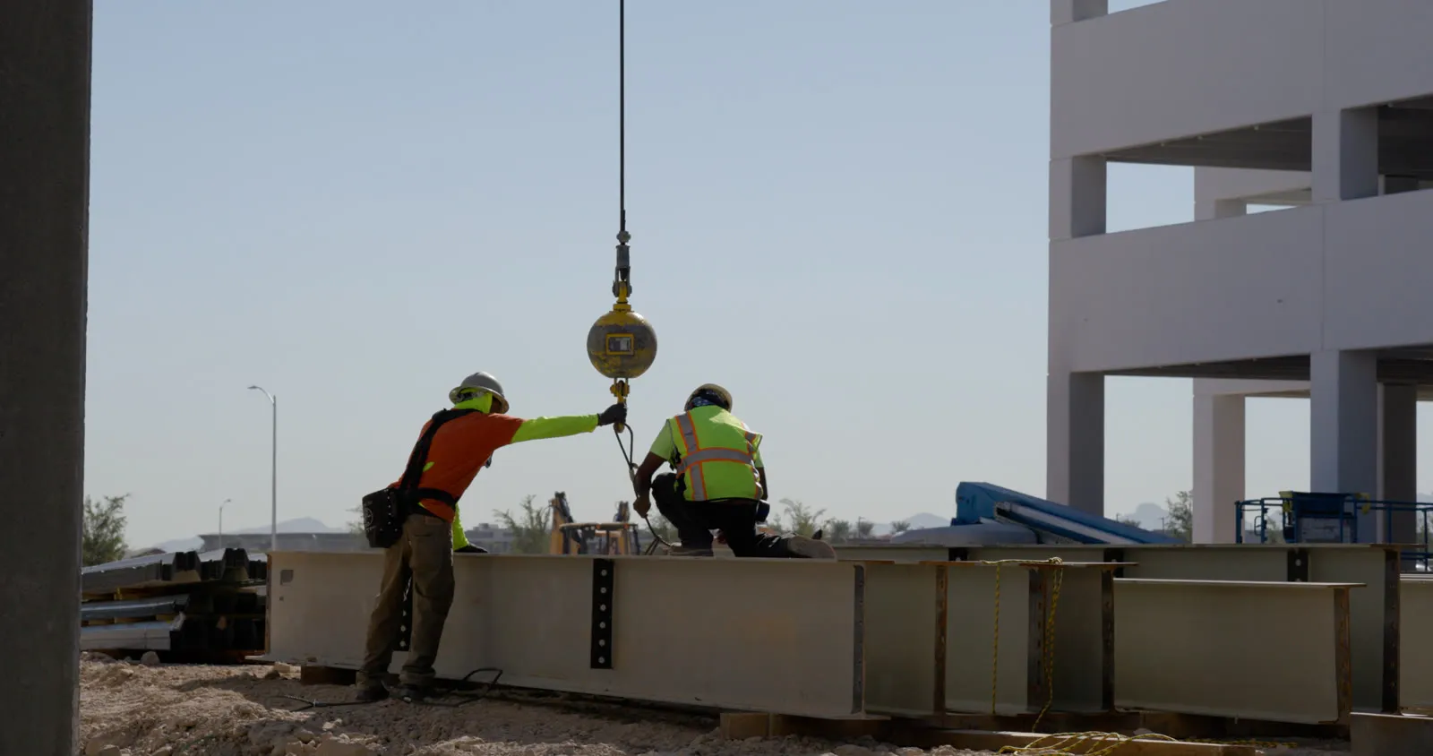 Two ironworkers in hard hats and hi-vis hooking a crane’s headache ball to a steel beam on the ground