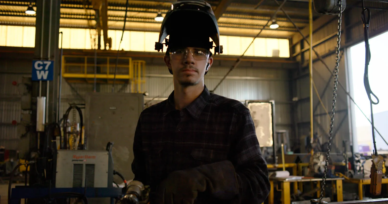 Young welder in a flannel shirt with his welding hood flipped up and safety glasses on, in a fabrication shop bay