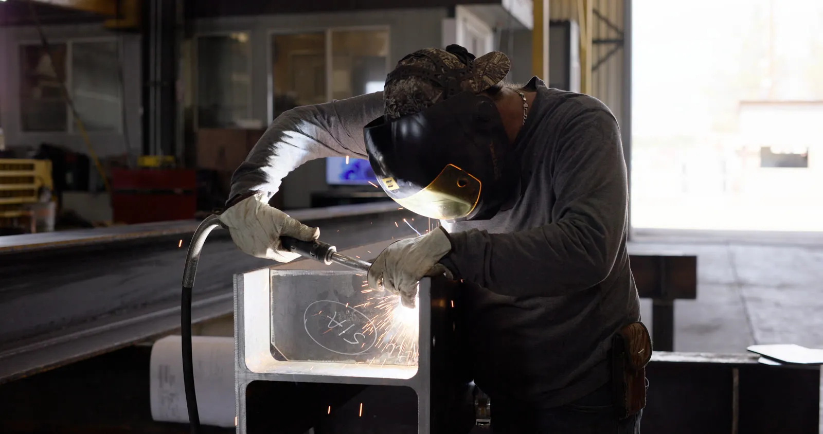 Fabricator welding a steel member in the shop in a welding helmet and leather gloves, sparks flying